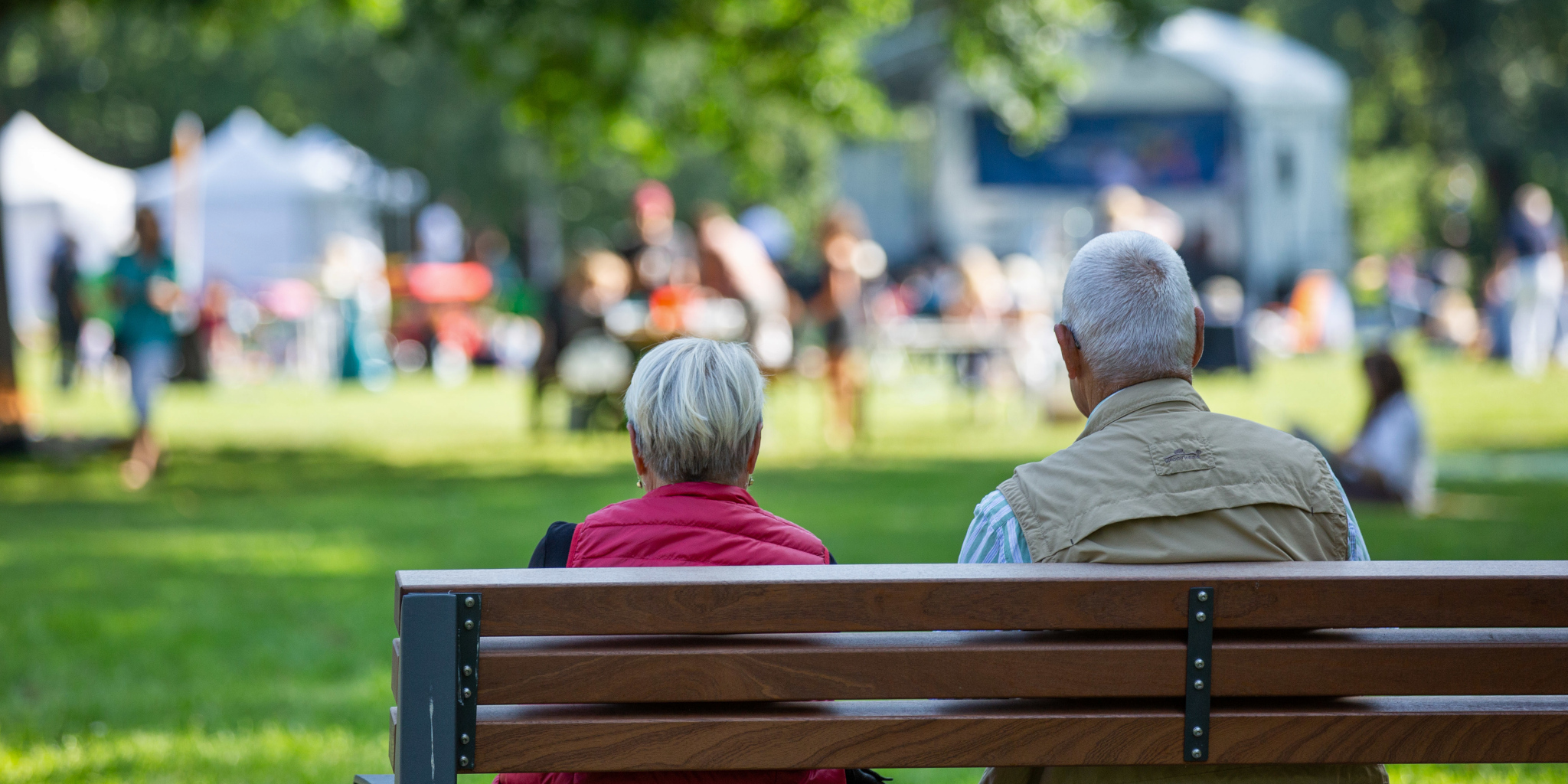 Eine ältere Frau und ein älterer Mann sitzen gemeinsam auf einer Bank und schauen auf eine Veranstaltung im Grünen.