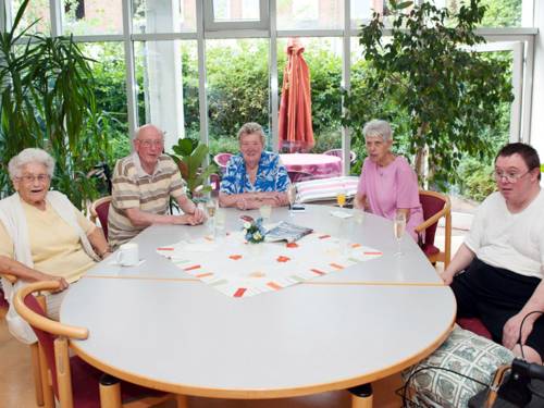 Fünf Bewohnerinnen und Bewohner einer Pflegeeinrichtung sitzen an einem großen Tisch eines Gemeinschaftsraums
