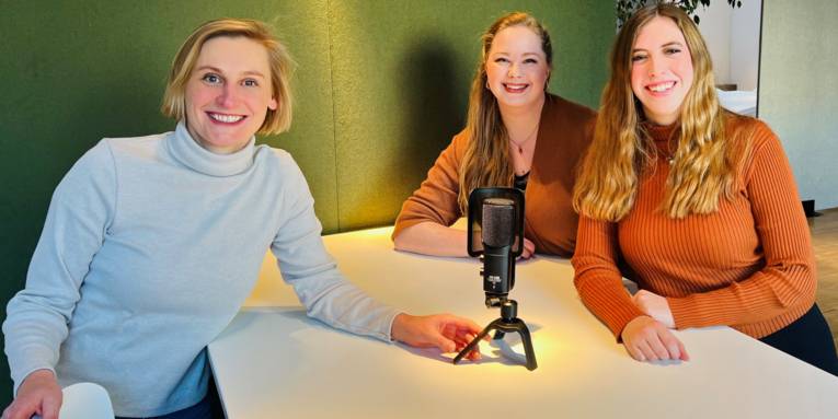 Drei Frauen sitzen an einem Tisch und schauen in die Kamera, auf dem Tisch steht ein Mikrofon.